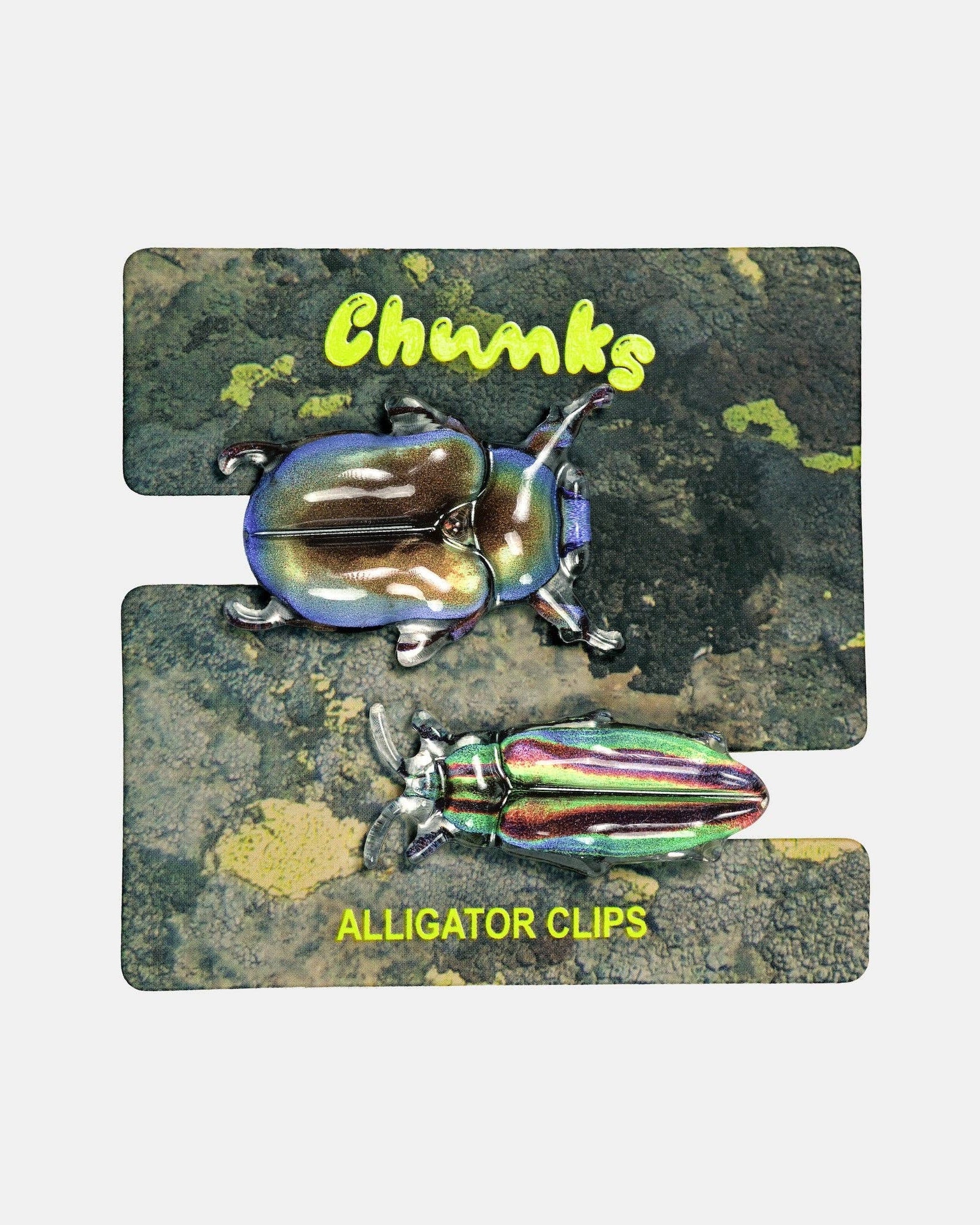A pair of iridescent alligator-style clips displayed on a printed backing card. The backing card has a dark textured background with two beetle illustrations and yellow text reading 'Chunks' and 'ALLIGATOR CLIPS'.