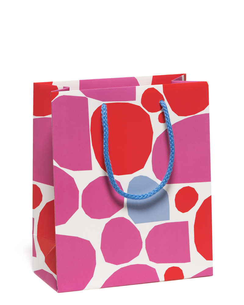 A paper shopping bag with a bold abstract pattern of pink, red, and magenta shapes on a white background, featuring blue rope handles.