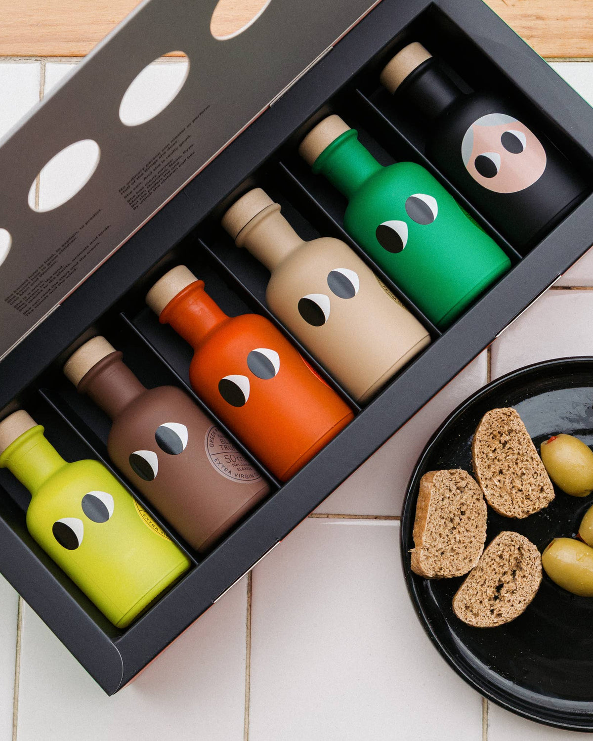A set of six wooden bottle-shaped objects in assorted colors arranged in a black gift box, photographed on a light tiled surface with a plate of olives and bread slices nearby.