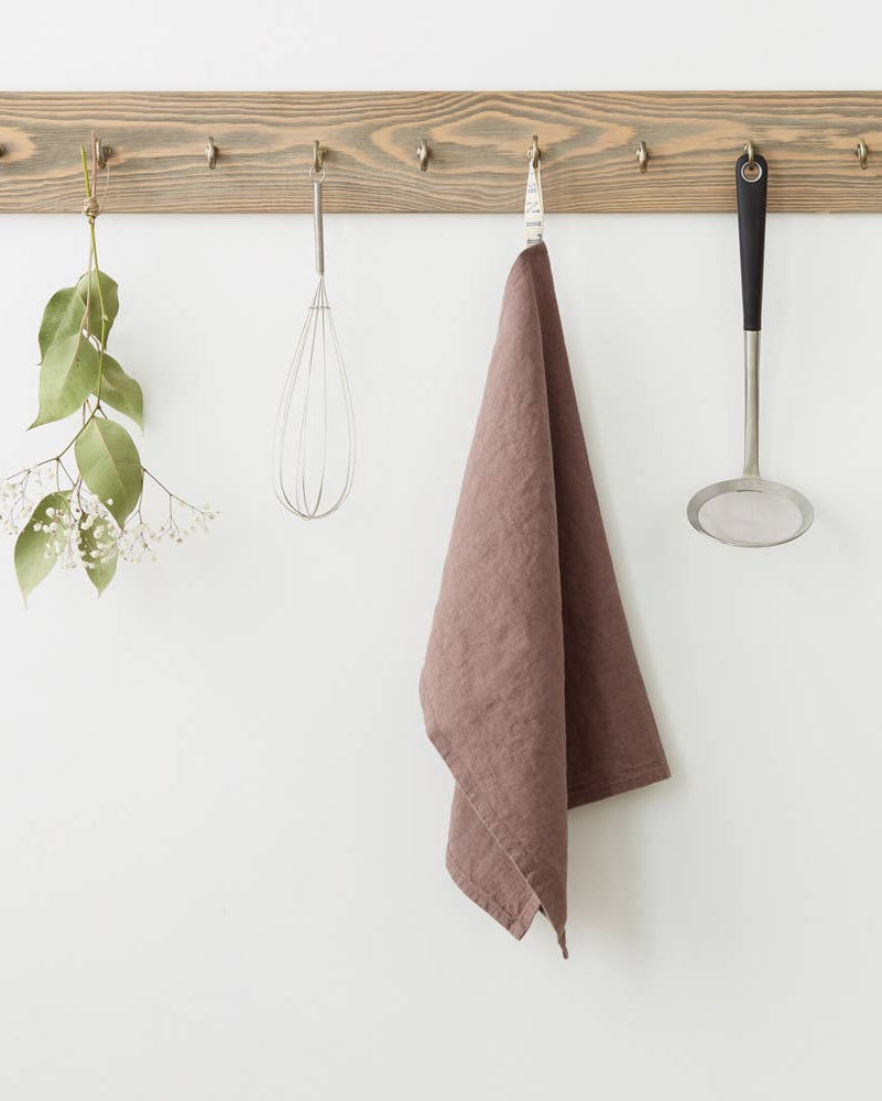 Wooden peg rack with hooks holding a brown towel, whisk, ladle, and green plant on a white wall.