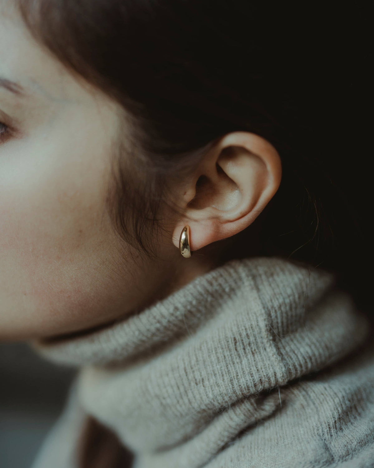 A close-up side profile of a person wearing a small gold crescent-shaped hoop earring, paired with a ribbed light gray turtleneck sweater.