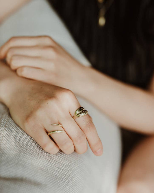A close-up lifestyle shot of a person's hand resting on a textured light gray surface, wearing three gold rings, including a distinctive double-loop ring on the index finger.