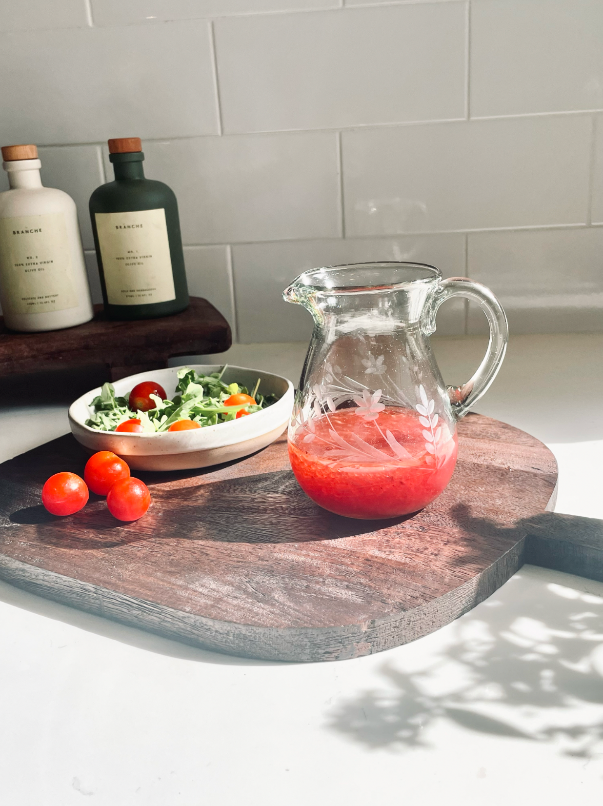 Glass pitcher with red liquid on a wooden cutting board next to a bowl of salad and tomatoes.