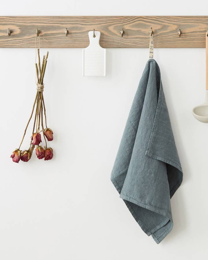 Wooden peg rail with hooks holding a blue towel, dried flowers, and a white container on a white wall.