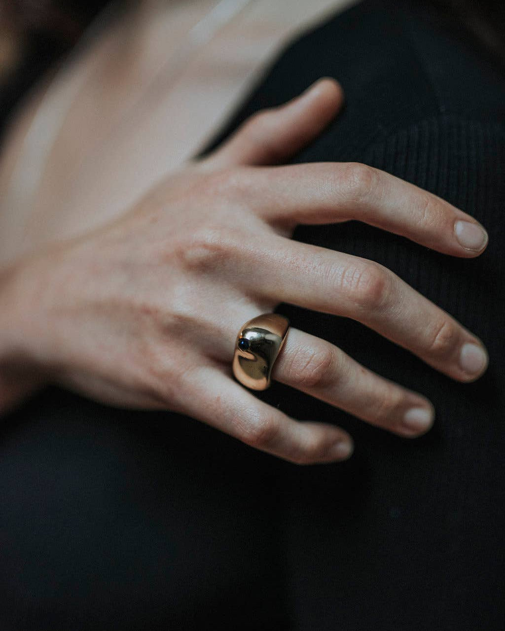 A close-up of a person's hand wearing a chunky, polished gold ring on one of the fingers, set against a dark ribbed top.