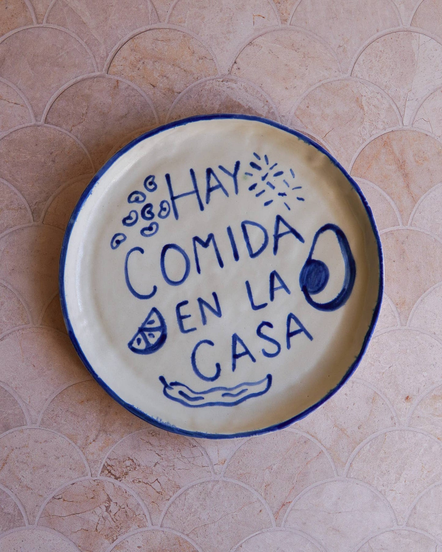 Ceramic plate with blue text 'Hay comida en la casa' on a tiled wall background