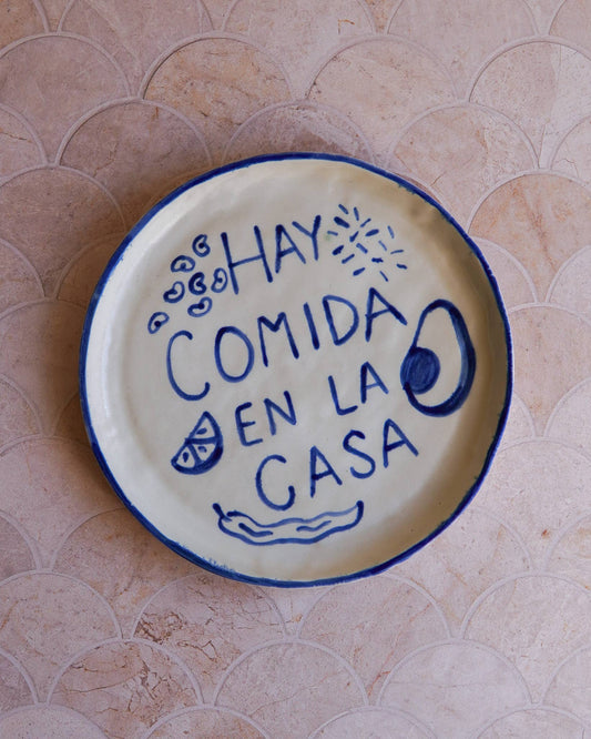 Ceramic plate with blue text 'Hay comida en la casa' on a tiled wall background