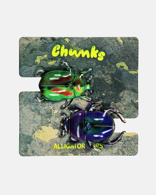 Two beetle-shaped jewelry charms on a split two-piece backing card. The top piece is metallic green with red and yellow accents, and the bottom piece is metallic purple with blue and pink accents.