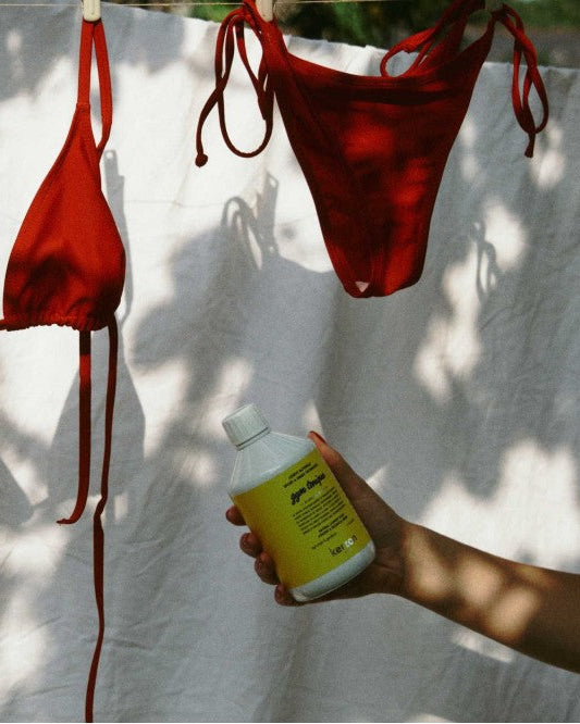 A hand holds a yellow bottle labeled 'sun block' in front of a white sheet with red bikini bottoms and a red bikini top hanging on a clothesline in a sunny, dappled outdoor setting.