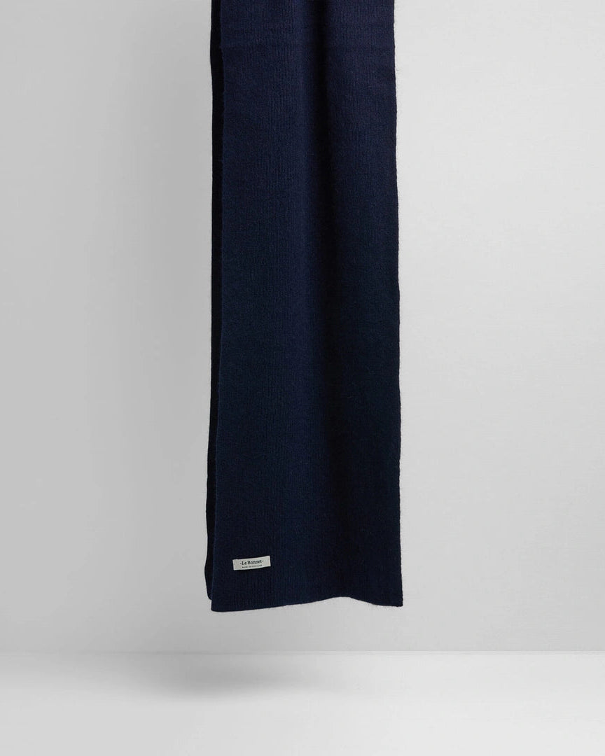 A dark navy blue knit scarf hanging vertically against a light gray studio background, with a small white brand tag near the bottom edge.
