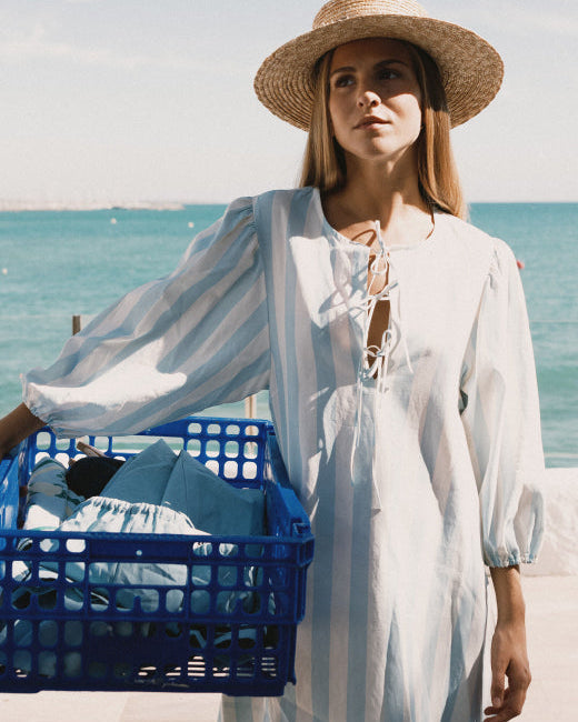 Woman in a white dress and straw hat holding a blue crate by the sea.