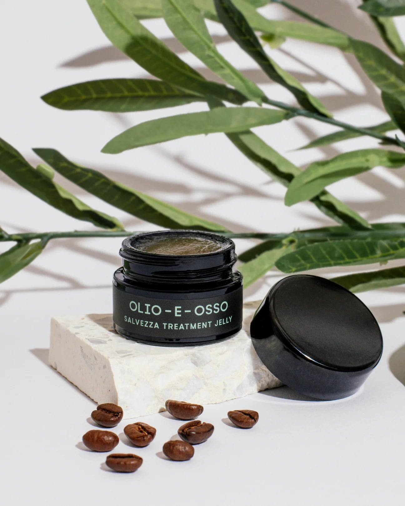Olio E Osso treatment jelly jar with coffee beans and green leaves on a light background