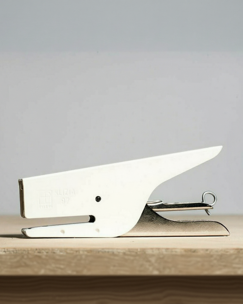 A white and silver desk stapler resting on a light wooden surface against a neutral gray background.