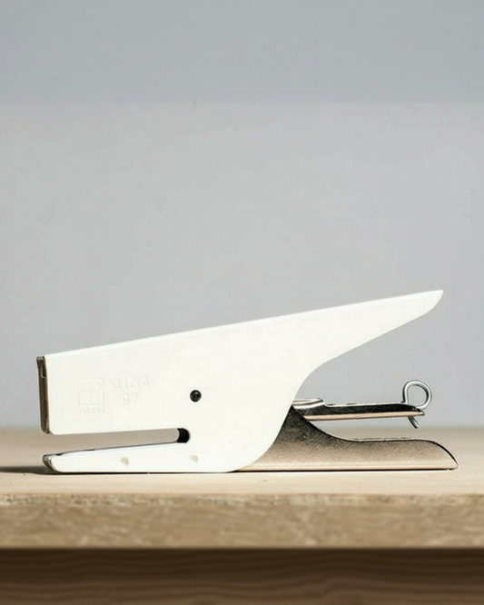 A white and silver desk stapler resting on a light wooden surface against a neutral gray background.